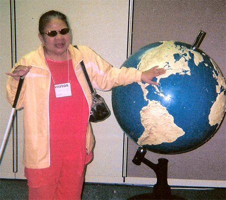 photo shows Nanta smiling and standing next to a world globe that is about 3 feet in diameter, mounted on a pedestal so the top is about 5 feet from the floor.  The land features of the globe, including mountains, are raised so they can be felt, and Nanta has her hand on the globe with her first fingertip on Maryland.